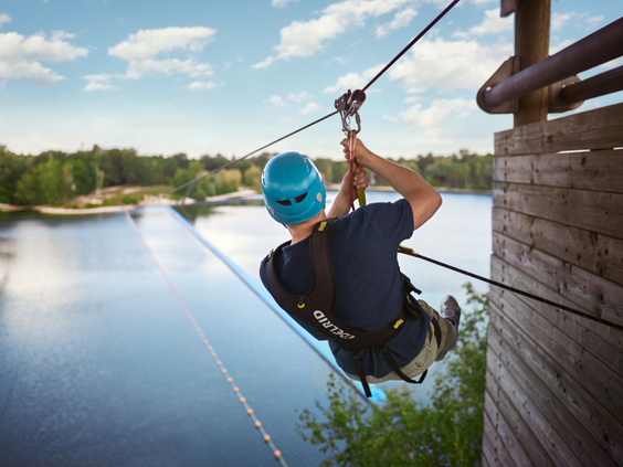 Een man met een helm en tuigje zoeft aan de zipline (tokkelbaan) over het grote meer van Center Parcs De Kempervennen.