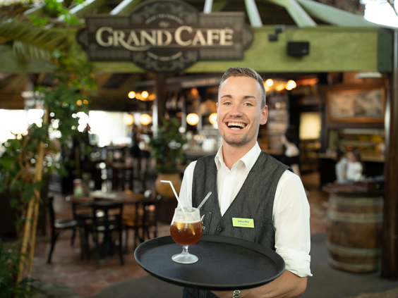 Een vriendelijke ober serveert een drankje op een dienblad voor de ingang van het sfeervolle Grand Café in de Market Dome van Het Meerdal.