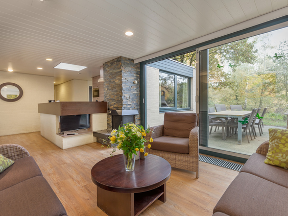 Overzicht van de ruime woonkamer in een cottage van Het Meerdal, met een zithoek, een open haard en grote ramen die uitkijken op de groene omgeving.