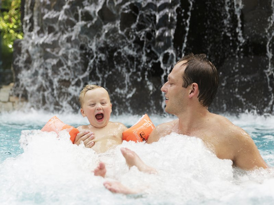 Een lachende vader en zoon genieten samen in het bruisende bubbelbad van de Aqua Mundo bij Het Meerdal, met op de achtergrond een grote waterval.