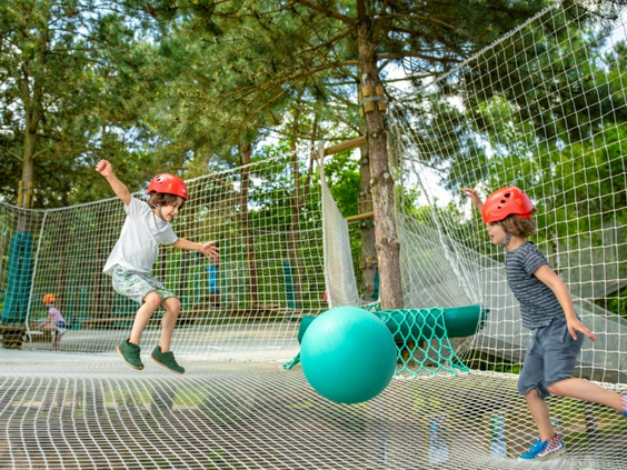 Twee jongens met rode helmen op spelen met een grote bal in een groot nettenparcours dat hoog tussen de bomen hangt in de indoor speelwereld.