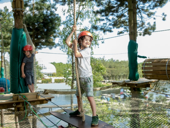 Een lachende jongen met een helm op houdt zich vast aan een touw terwijl hij balanceert op het High Adventure Experience klimparcours van Het Meerdal.