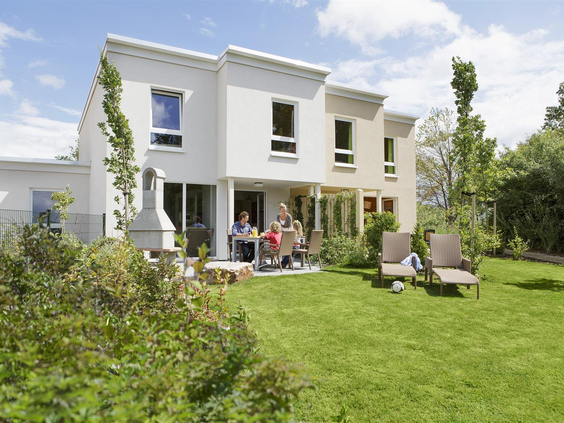 De buitenkant van een moderne witte cottage met een gezin dat ontspant op het terras in de ruime, groene tuin bij Center Parcs Park Bostalsee.