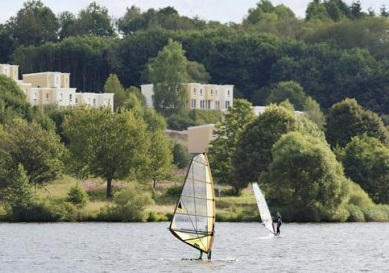 Een windsurfer op het water van het Bostalmeer, met op de achtergrond de vakantiehuizen van Center Parcs Park Bostalsee.
