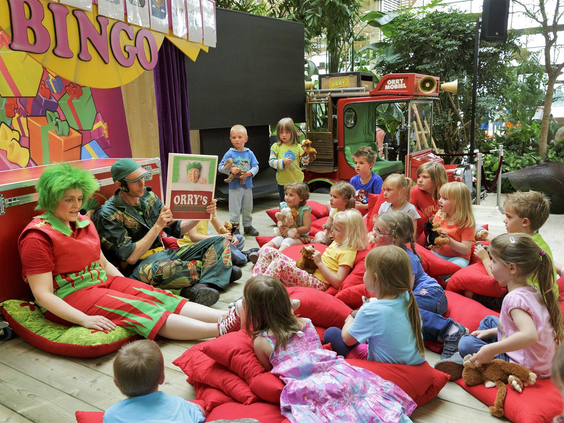 Een groep kinderen zit op rode kussens te kijken naar een voorstelling met poppen en acteurs in de Kids Club van Center Parcs Park Bostalsee.