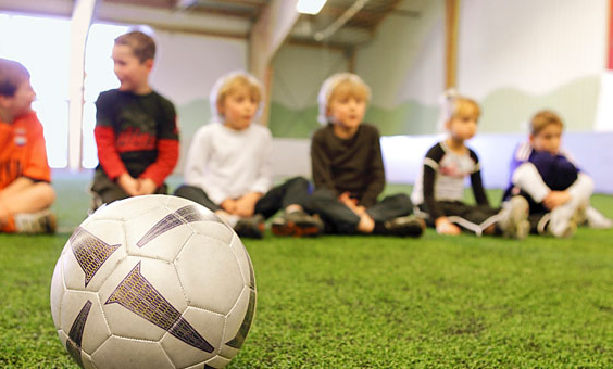 Kinderen luisteren naar de coach tijdens een voetbalclinic in de sporthal van Center Parcs Park Hochsauerland, met een voetbal op de voorgrond.