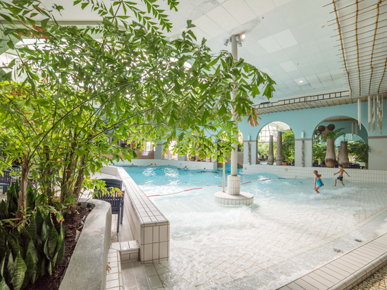 Kinderen spelen in de golven van het golfslagbad in de tropische Aqua Mundo van Center Parcs Port Zélande, omgeven door palmbomen.