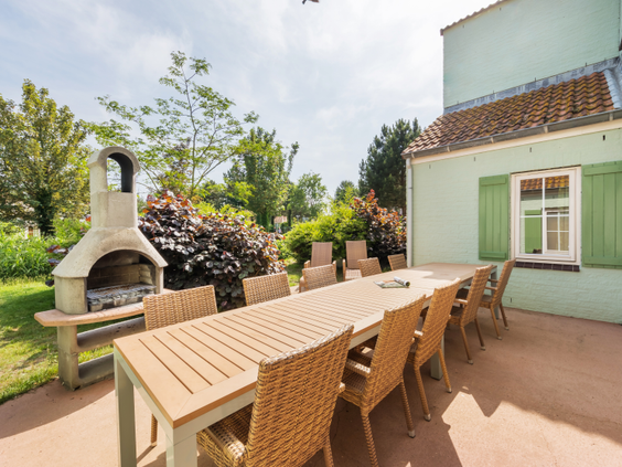 Een groot terras van een vakantiehuis op Center Parcs Port Zélande met een lange houten eettafel en een stenen barbecue.