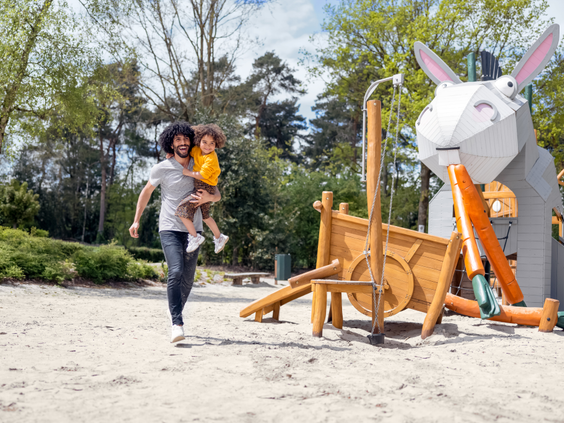 Een vader rent met zijn kind in de armen door een zandspeeltuin bij Center Parcs Erperheide, met een groot speeltoestel in de vorm van een konijn.
