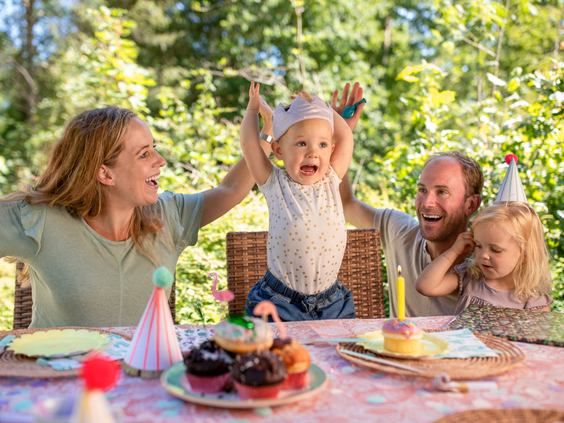 Een gezin met twee jonge kinderen viert een verjaardag aan een versierde picknicktafel in het bos, met cupcakes en feestmutsen, bij Center Parcs Erperheide.