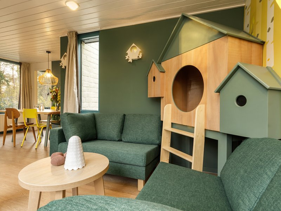 De woonkamer van de 'Dieren in het bos' themacottage bij Erperheide, met een groene hoekbank en een ingebouwd houten speel- en slaaphuisje in de vorm van vogelhuisjes.