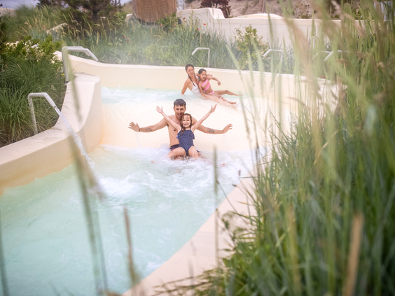 Een gezin glijdt met veel plezier samen van de brede familieglijbaan in het buitenbadgedeelte, omgeven door wuivend duingras bij Center Parcs Zandvoort.