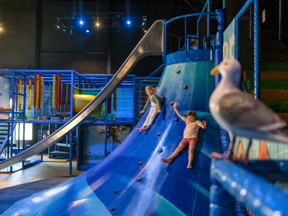 Kinderen klimmen en klauteren op de blauwe speeltoestellen met een glijbaan in de Beach Factory indoor speeltuin van Center Parcs Zandvoort.