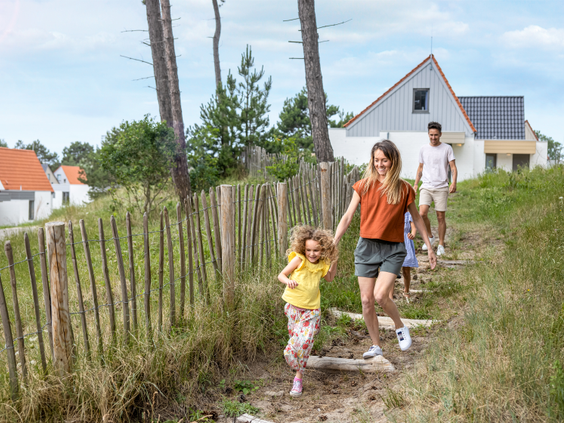 Een gezin wandelt hand in hand over een pad door het duinlandschap op vakantiepark Center Parcs Zandvoort, met een cottage op de achtergrond.