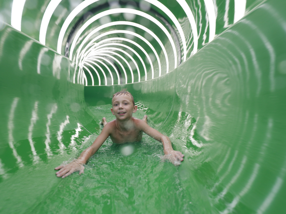 Een jongen glijdt met een grote glimlach door een groene, verlichte waterglijbaantunnel in de Aqua Mundo van Center Parcs Zandvoort.