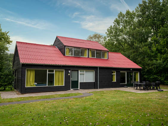 Een vrijstaande, zwarte bungalow met een rood dak en een grote tuin op Villapark de Bloemert, omgeven door bomen.