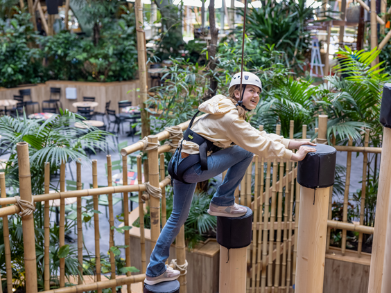 Een gezekerd meisje met een helm op klimt over een hindernis op het indoor klimparcours Via Ferrata, hoog boven de Market Square van De Eemhof.