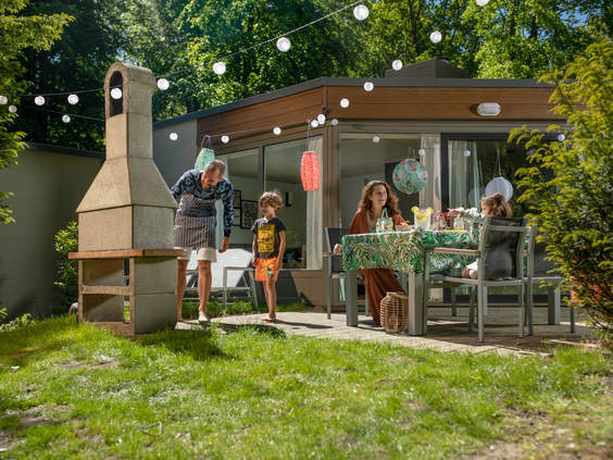 Een gezin geniet samen van een gezellige barbecue op het zonnige terras van hun cottage bij Center Parcs De Eemhof, met sfeervolle lampionnen en lampjes.