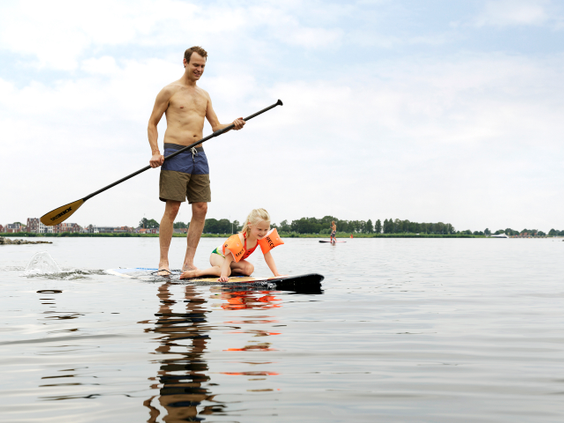 Een vader staat met een peddel op een sup-board terwijl zijn dochter voorop zit, op het grote Eemmeer bij Center Parcs De Eemhof.