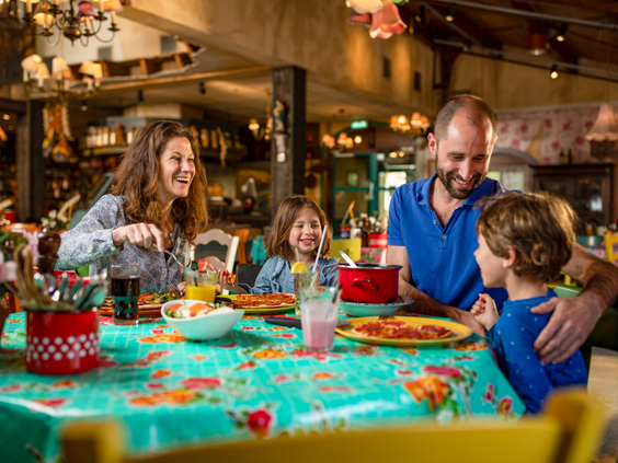 Een lachend gezin met twee kinderen geniet van een gezellige maaltijd aan een kleurrijk gedekte tafel in een van de restaurants in de Market Square van De Eemhof.