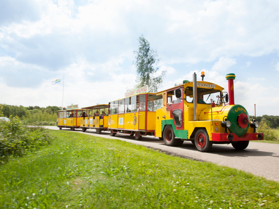 De vrolijk gekleurde gele parktrein van Molecaten Park De Flaasbloem maakt een rondrit door het groene landschap van de camping.