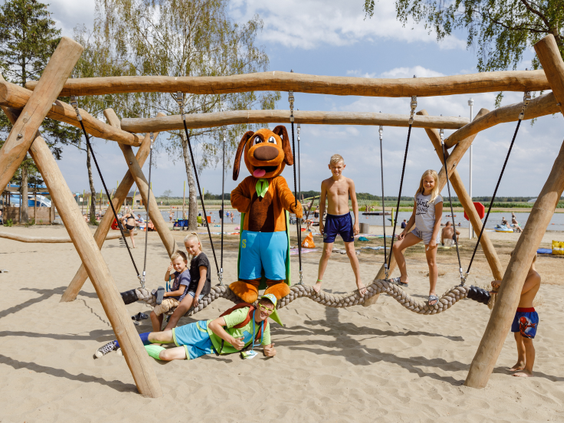 Kinderen spelen samen met de Molecaten-mascotte op een houten klautertoestel op het zandstrand van Molecaten Park De Flaasbloem.