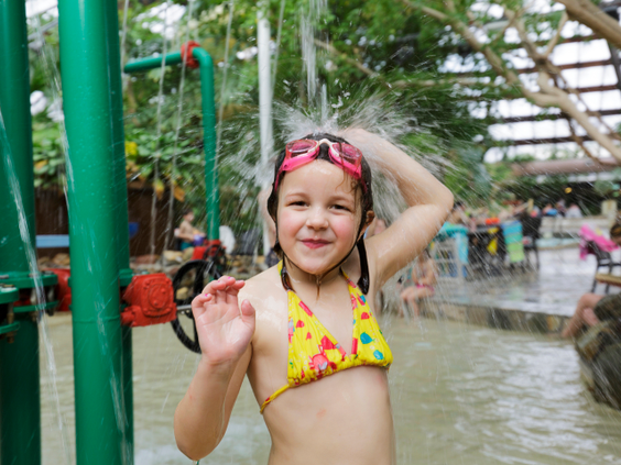 Een lachend meisje in bikini en met een zwembril op haar hoofd zwaait naar de camera terwijl ze onder een waterdouche in de Aqua Mundo staat.
