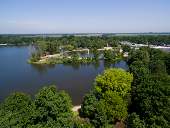 Een luchtfoto van Center Parcs De Huttenheugte op een zonnige dag, met uitzicht over het grote meer, het strandje, de cottages en de omliggende bossen.
