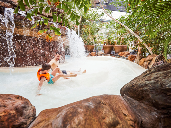 Een vader en zijn zoon met zwemvleugels glijden samen door de wildwaterbaan van de Aqua Mundo bij Center Parcs De Huttenheugte, langs rotsen en watervallen.
