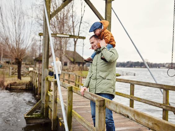 Een vader met zijn zoontje op de schouders wandelt over een houten brug bij het meer van Center Parcs De Huttenheugte.