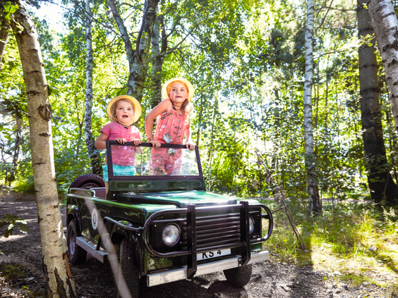 Twee vrolijke meisjes in roze kleding staan in een elektrische kinder-jeep en rijden door het bos tijdens de Kids Safari bij Center Parcs De Huttenheugte.