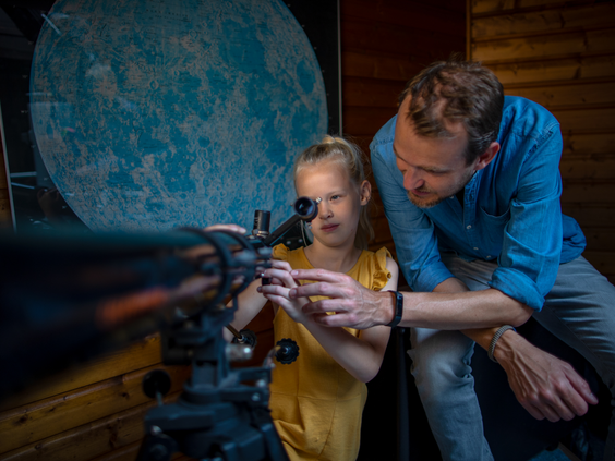 Een vader helpt zijn dochter met het kijken door een telescoop naar een grote afbeelding van de maan op het Avontureneiland van Center Parcs De Huttenheugte.