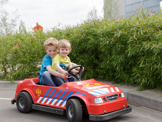 Twee blije jongens rijden samen in een rode speelgoed-brandweerauto door het Kids Verkeerspark bij Center Parcs De Huttenheugte.