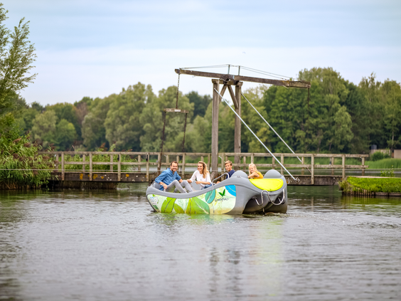 Een gezin met twee kinderen geniet van een tochtje in een bootje op het grote meer bij Center Parcs De Huttenheugte, met een ophaalbrug op de achtergrond.