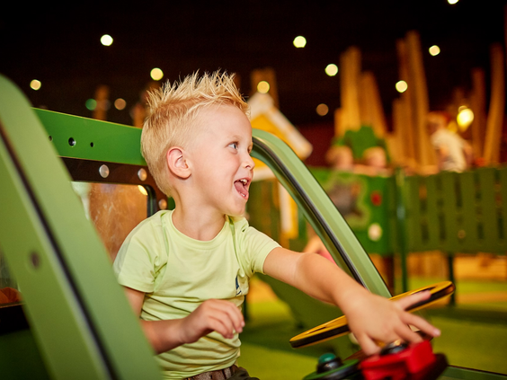Een lachende jongen met blond haar zit in een groen speeltoestel en drukt op een knop in de indoor speeltuin La Selva op De Leistert.