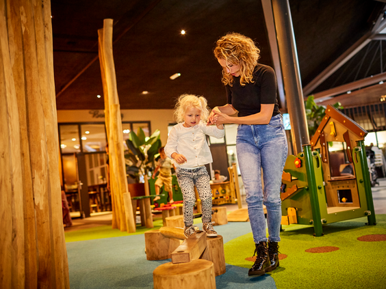 Een moeder helpt haar jonge dochter met balanceren op houten paaltjes in het peutergedeelte van de indoor speeltuin op Recreatiepark De Leistert.