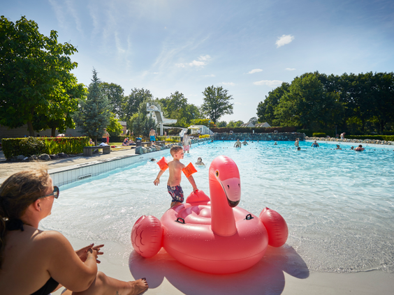Een kind speelt met een opblaasbare roze flamingo in het buitengolfslagbad van Recreatiepark De Leistert op een zonnige dag.