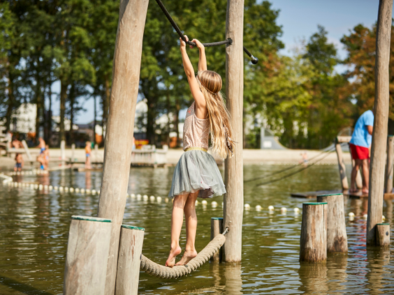 Een meisje met lang haar balanceert op een touwparcours boven het water in de avontuurlijke waterspeeltuin van De Leistert.