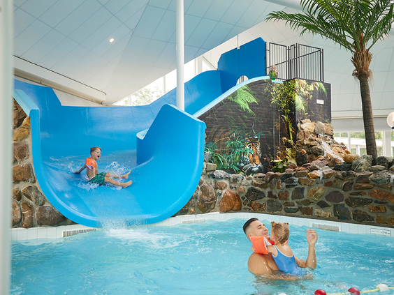 Een peuter glijdt van een brede blauwe glijbaan het kinderbad in van het subtropische zwembad op Recreatiepark De Leistert, waar een vader hem opwacht.