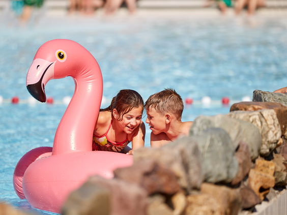 Twee kinderen fluisteren geheimpjes terwijl ze in het zwembad dobberen met een grote, roze, opblaasbare flamingo.