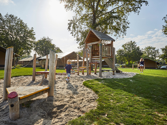 Een houten speeltoestel met glijbaan in een zandspeeltuin op Recreatiepark De Leistert, omgeven door een groot grasveld en bomen.