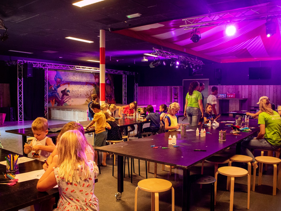Een groep kinderen zit aan lange tafels te knutselen tijdens een activiteit van het animatieteam in de met paars licht verlichte theaterzaal van De Leistert.