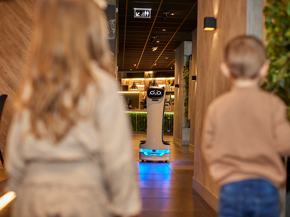 Twee kinderen kijken gefascineerd naar een serveerrobot die met blauwe lichten door de gang van een restaurant op De Leistert rijdt.