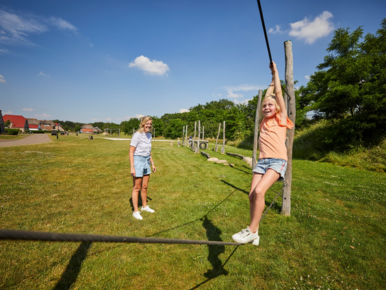Een meisje zoeft aan de kabelbaan in de buitenspeeltuin van Recreatiepark De Leistert, terwijl een vriendinnetje kijkt.