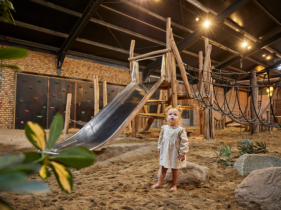Een peuter staat in het zand van de indoor speeltuin La Selva op Recreatiepark De Leistert, naast een metalen glijbaan.