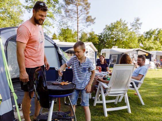 Een gezellig gezin dat aan het barbecueën is bij hun kampeerplek op RCN de Roggeberg, waarbij de zoon de vader helpt bij de BBQ.