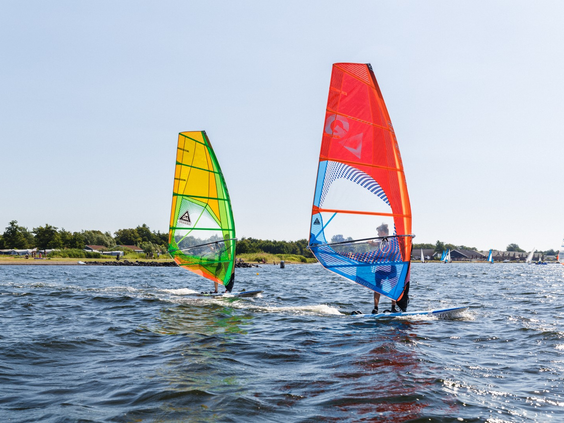 Twee windsurfers met kleurrijke zeilen varen over het water van het Veerse Meer bij de surfschool van RCN de Schotsman.