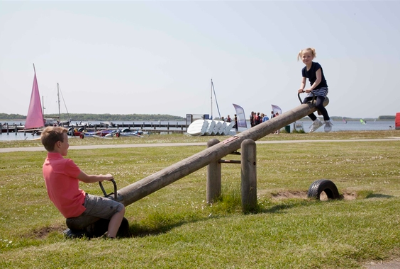 Twee kinderen spelen samen op een houten wipwap op een grasveld aan het water bij RCN de Schotsman, met zeilbootjes op de achtergrond.