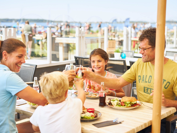 Een vrolijk gezin proost met hun drankjes op het terras van de brasserie bij RCN de Schotsman, met uitzicht op het water.