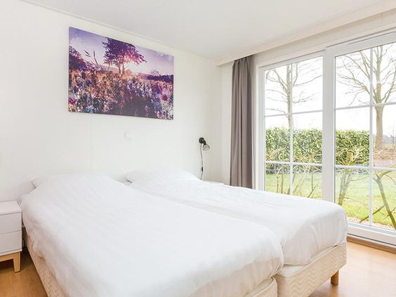 Lichte slaapkamer in een vakantiehuis op De Tolplas, met een opgemaakt tweepersoonsbed en grote ramen die uitkijken op de groene tuin.